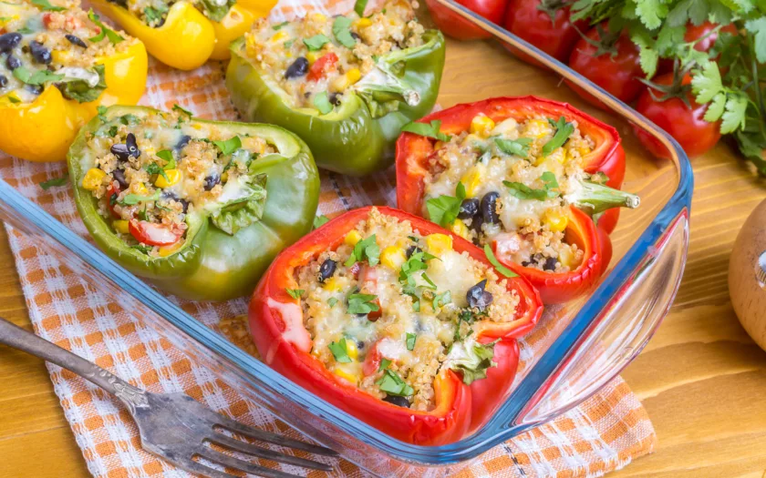 Vibrant and colorful stuffed bell peppers filled with a mix of quinoa, black beans, corn, and melted cheese, garnished with fresh cilantro. The peppers, in shades of red and green, sit neatly in a glass baking dish atop a wooden table with a checkered cloth, ready to be enjoyed